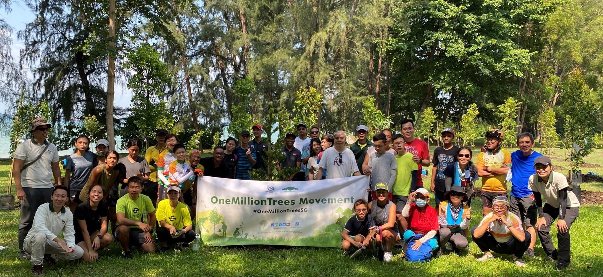 A group is gathered outside holding a banner that reads "OneMillionTrees Movement #OneMillionTreesSG" among trees.